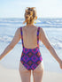 Bold patterned one-piece swimsuit with retro evil eye floral design, styled in festival-ready outdoor settings. Swimsuit features a scoop neckline, cheeky fit, and low scoop back. Ideal for summer vacations, pool parties, raves, and alt girl fashion. Made from stretchy, moisture-wicking fabric with UPF 50+ protection. Available in sizes XS to 3X. Colorful, statement swimwear designed for confident, maximalist style lovers.